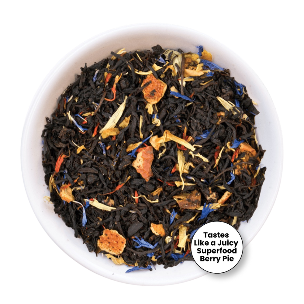 A top-down view of a white bowl filled with loose-leaf herbal tea blend. The tea contains dark dried leaves mixed with orange dried fruit pieces, yellow dried flowers, and small blue accents.