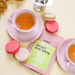A box of Sweet Macaron tea next to a cup of tea and pink macarons on a plate.