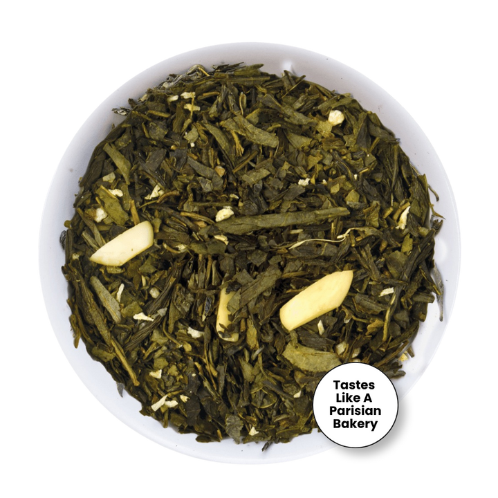 Loose leaf tea in a white bowl, featuring dark green tea leaves with two pale garlic cloves and small white flower petals. A white circular badge in the bottom right corner reads &