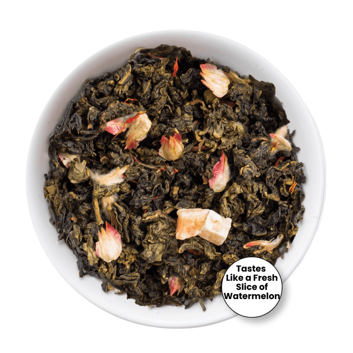 A top-down view of loose-leaf tea in a white bowl, featuring dark green tea leaves mixed with pale pink watermelon blossom pieces. A circular badge in the bottom right corner reads, &