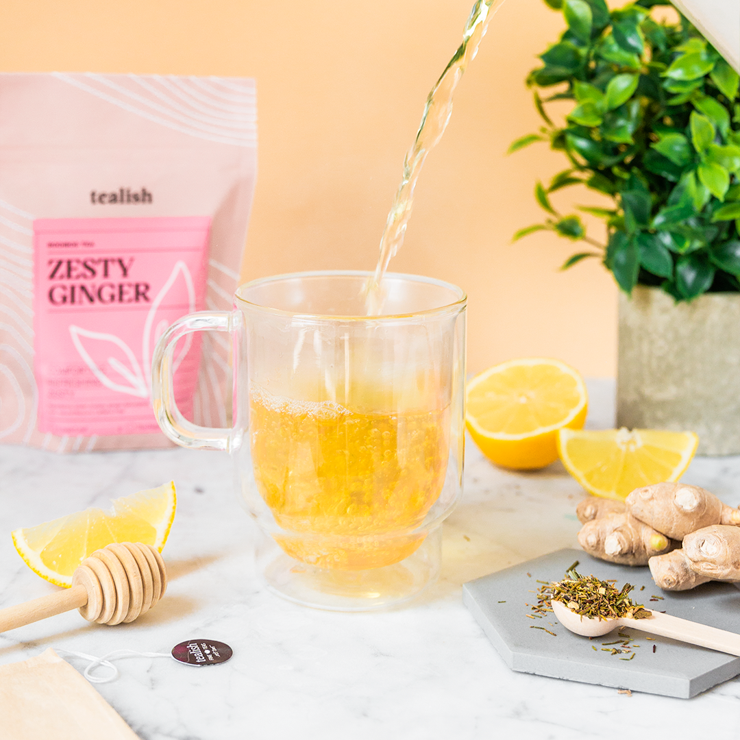 A cup of tea being prepared with loose leaf tea from a bag labeled 'Zesty Ginger'. Ingredients such as ginger, lemon, and honey are visible, along with a 'Tealish' brand tag.