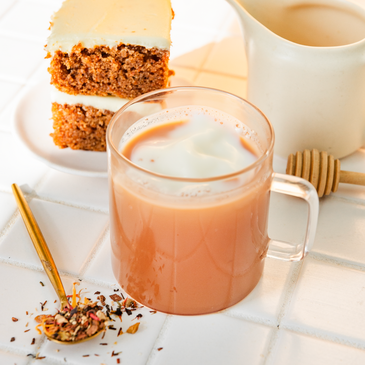 A cup of carrot cake flavored rooibos tea with a slice of carrot cake, a spoon with loose tea leaves, and a honey dipper on a white surface.