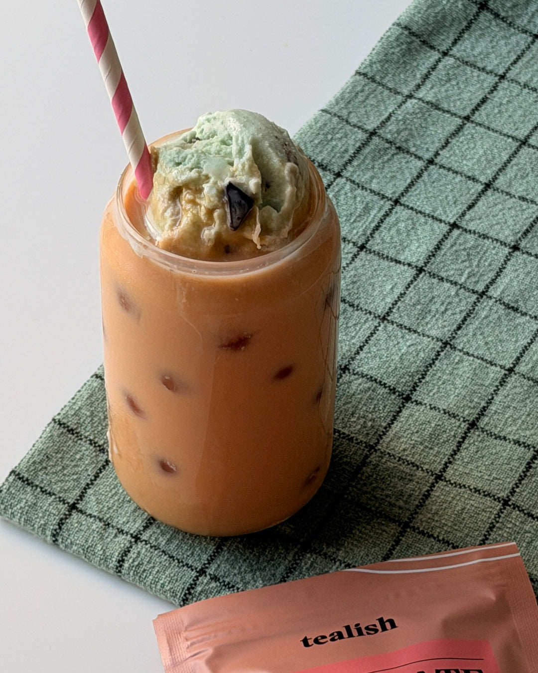 Milkshake with ice cream and a straw on a checkered cloth, next to a Tealish Chocolate Mint tea package.