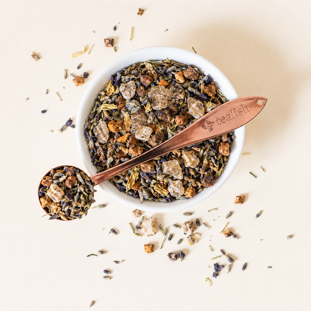 A top-down view of a white bowl filled with a loose herbal tea blend, with a copper spoon resting across the bowl and a few pieces scattered around on a light beige background. The spoon is engraved with the brand name 'tea fish'.