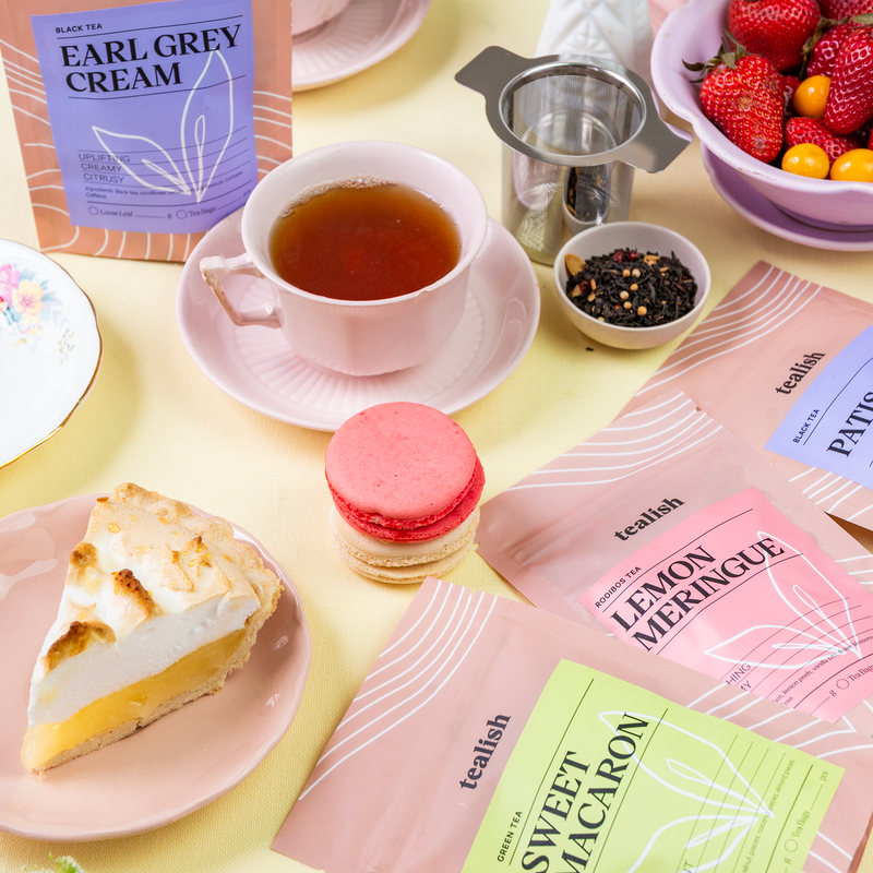 An assortment of dessert-inspired loose leaf teas from tealish, displayed with tea-making accessories and desserts on a table. The image shows teacups, a teapot, tea filters, strawberries, a lemon, and a piece of cake.