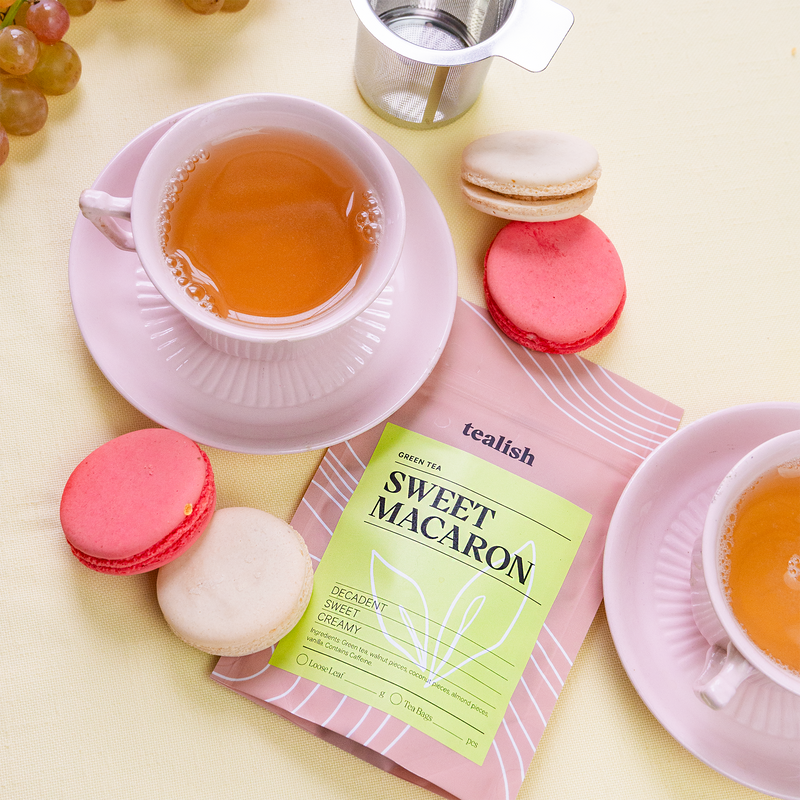 A box of Sweet Macaron tea next to a cup of tea and pink macarons on a plate.