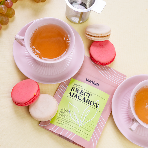 A box of Sweet Macaron tea next to a cup of tea and pink macarons on a plate.