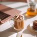 Cup of tea latte with whipped cream and chocolate shavings on a desk with a laptop and cookies.