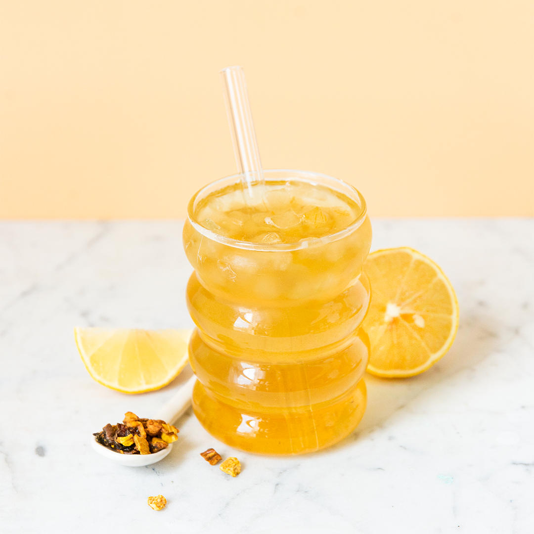 A glass of citrusy looking iced tea with a straw, accompanied by lemon slices and what appears to be loose tea leaves on the side.