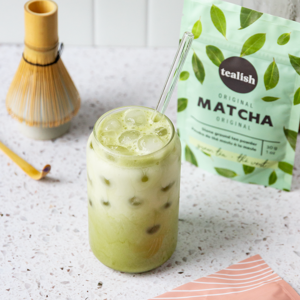 A glass of matcha green tea with ice and a straw, alongside a package of Tealish Original Matcha powder and a bamboo whisk.
