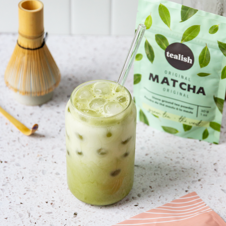 A glass of matcha green tea with ice and a straw, alongside a package of Tealish Original Matcha powder and a bamboo whisk.