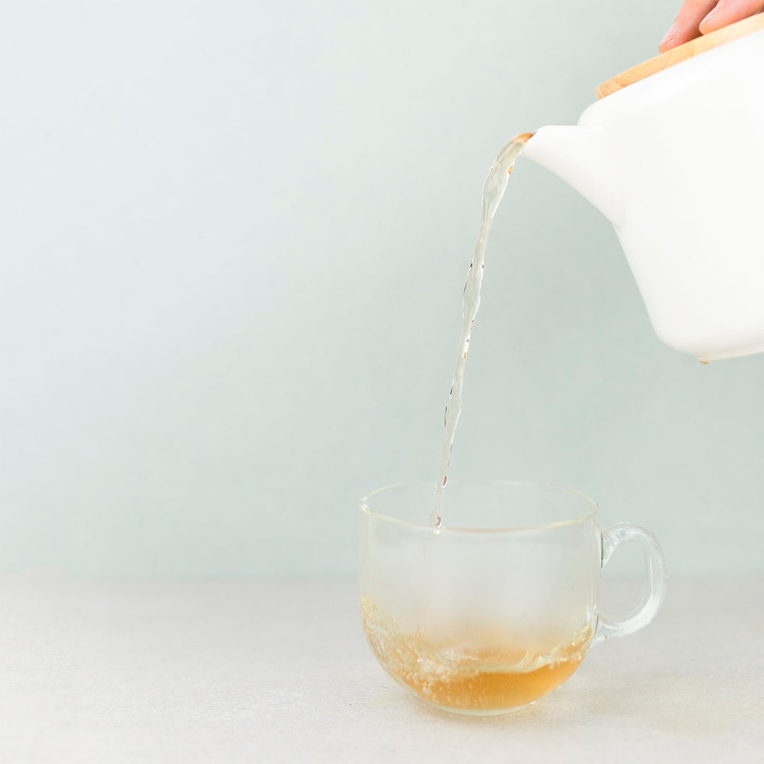 A cup of tea being poured from a white teapot into a clear glass mug on a light background.