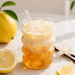 Glass of iced lemonade with a straw, surrounded by lemons and flowers on a light surface.