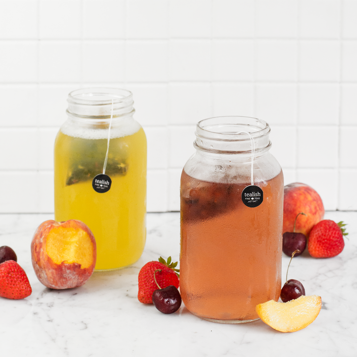 Two glass jars on a kitchen countertop filled with peach-colored liquid, presumably peach jasmine tea, with ice cubes. Fresh peach slices, strawberries, and cherries are scattered around, indicating the flavor of the tea.
