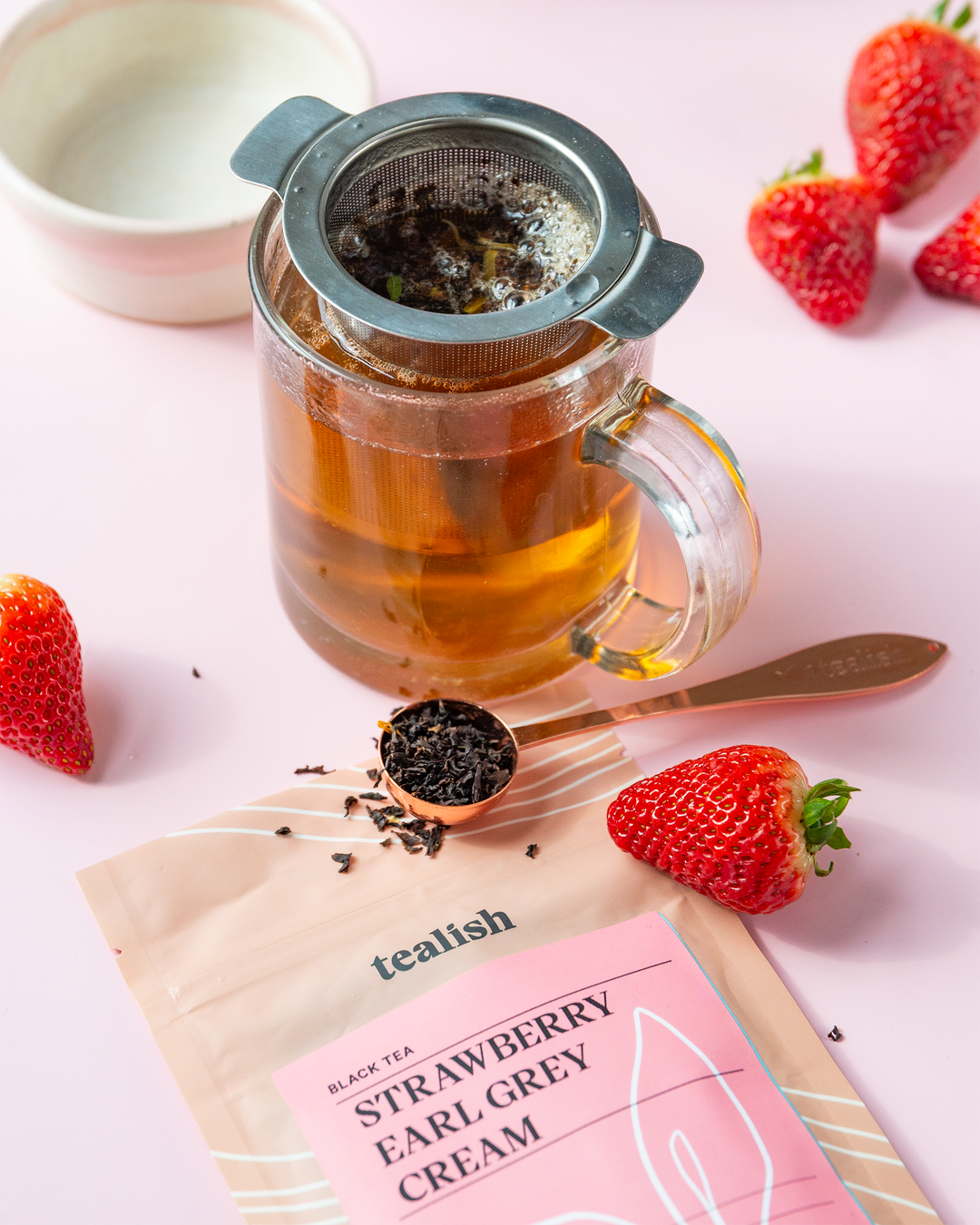 A tea bag labeled Strawberry Earl Grey Cream is steeped in a cup, surrounded by fresh strawberries on a pink background. A spoon with tea leaves on it adds a tactile element to the visual.