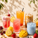 Four colorful iced drinks in clear glasses arranged on a tabletop with fresh fruit and herbs around them. The drinks include a pink strawberry iced milk, an orange citrus drink, an iced coffee, and a light blue milk-based beverage.