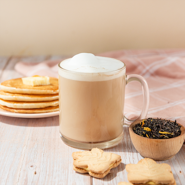A cup of sweet Canadian maple black tea with a frothy top, alongside a small bowl of maple syrup and dried petals, with cookies and a stack of pancakes in the background.