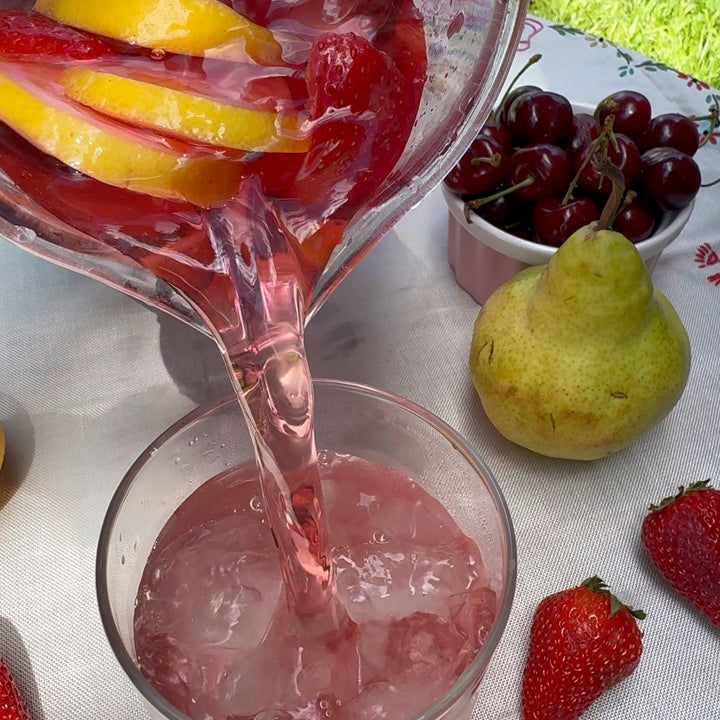 Pink drink being poured from a pitcher into a glass with fruit on a table outdoors.