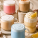 Five colorful iced coffee drinks in clear glass mugs with handles, arranged on a beige surface with shadows, whipped cream, lemon slices, vanilla beans, and coconut pieces. The drinks have pink, tan, light blue, and yellow colors with thick foam tops.
