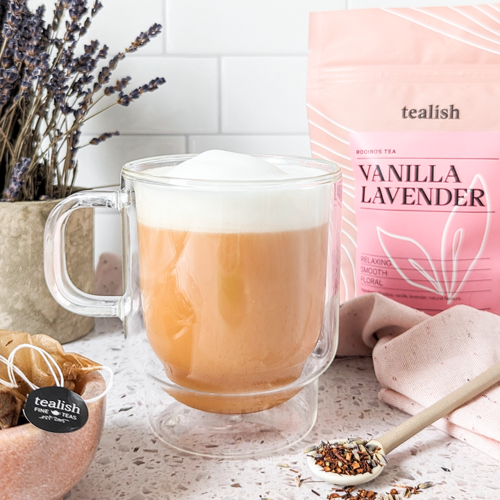A clear glass mug filled with a creamy beverage, alongside a box of TeaLish Vanilla Lavender tea, with dried lavender and a spoon with tea leaves scattered around.
