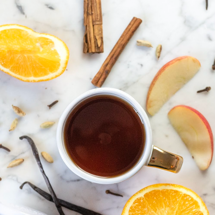 A cup of black tea with pieces of orange and apple, and cinnamon sticks around it on a marble surface.