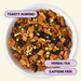 A top-down view of a white bowl filled with loose herbal tea blend labeled 'TOASTY ALMOND,' with additional text badges reading 'HERBAL TEA' and 'CAFFEINE FREE' on a light cream background.